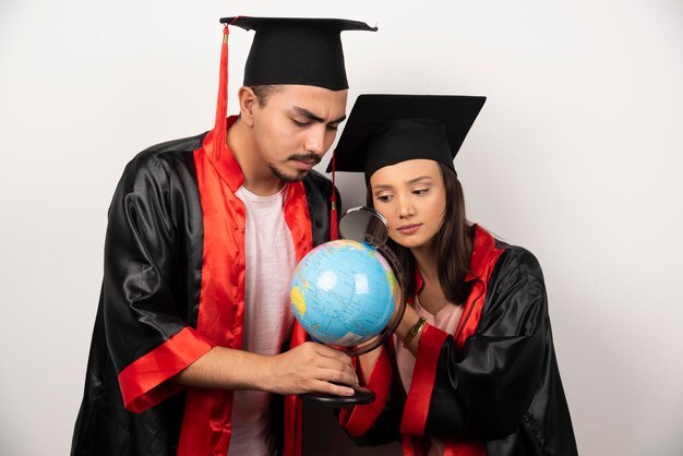 Pair of students in gown looking at globe with magnifying glass.