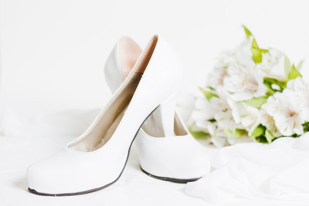 Pair of white high heels with scarf and flower bouquet on white backdrop