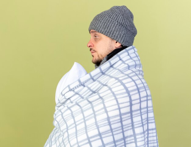 Бледный молодой блондин больной мужчина в зимней шапке и шарфе, завернутый в плед, стоит боком, держа подушку, изолированную на оливково-зеленой стене