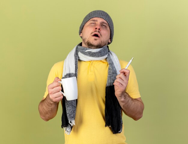 겨울 모자와 스카프를 착용 창백한 젊은 금발의 아픈 슬라브 남자는 컵과 온도계 복사 공간이 올리브 녹색 벽에 고립 된 보유