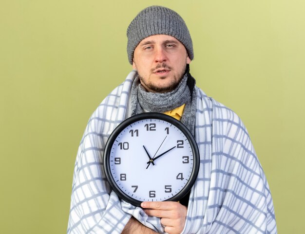 Бледный молодой блондин больной славянский мужчина в зимней шапке и шарфе, завернутый в плед, держит часы, изолированные на оливково-зеленой стене с копией пространства