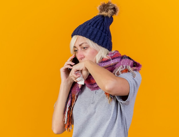 冬の帽子とスカーフを身に着けている淡い若い金髪の病気のスラブ女性が電話で話し、コピースペースでオレンジ色の壁に隔離された組織を保持します