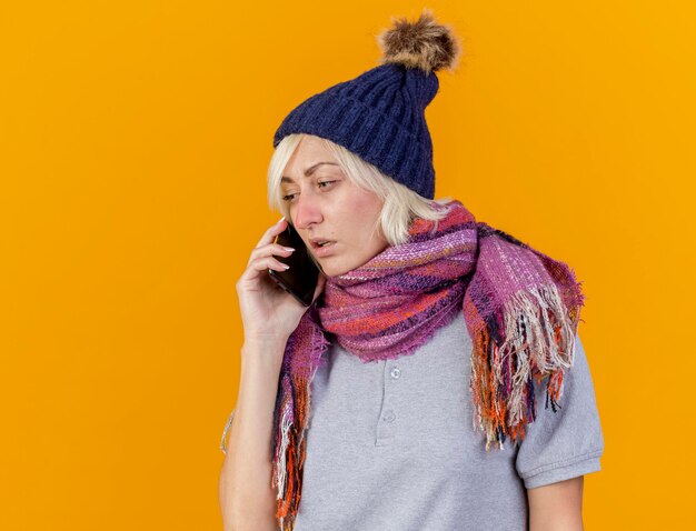 Бледная молодая блондинка больная славянская женщина в зимней шапке и шарфе разговаривает по телефону, изолированном на оранжевом