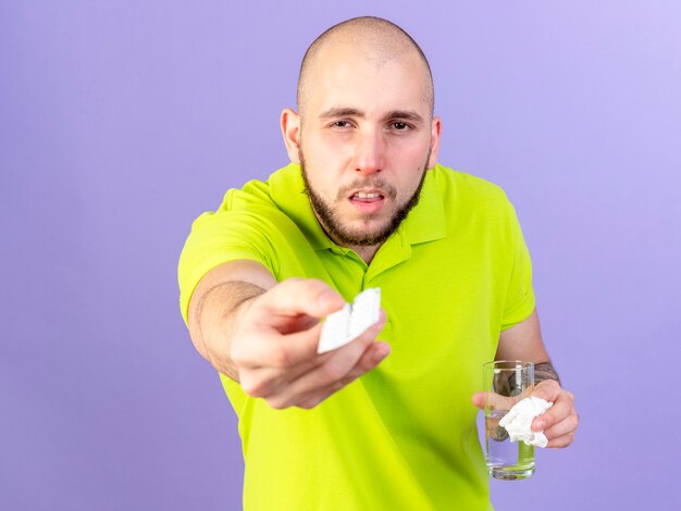 창백한 젊은 백인 아픈 남자는 보라색에 의료 약 팩과 물 한잔을 보유하고 있습니다.