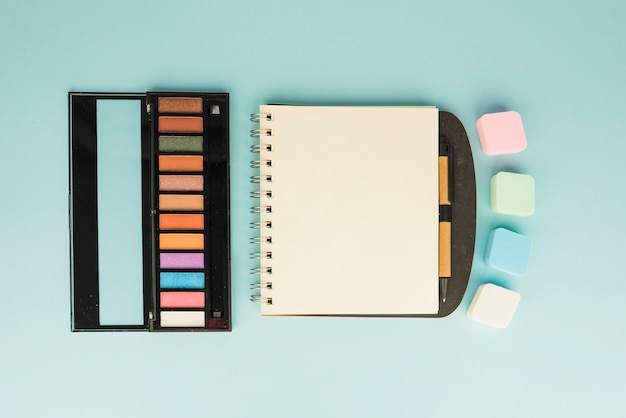 Palette of make up with colorful sponge and blank spiral notepad on colored backdrop