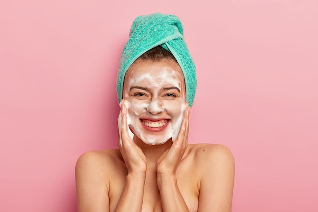 Free photo pampering and hygiene concept. happy young european woman massages cheeks, aplies bubble foam, washes face, smiles positively, has naked body, enjoys taking shower, wants to have clean skin.