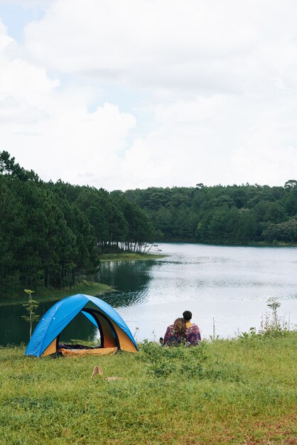 A pan shot of couple cuddling at the river near the tent with their backs to the camera