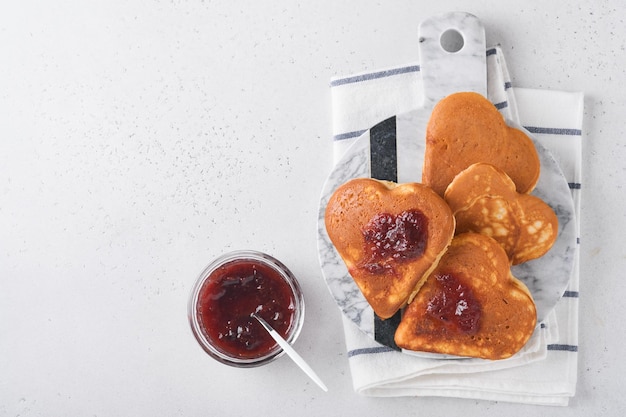베리 잼과 꿀을 하트 모양으로 만든 팬케이크와 흰색 나무 배경 위에 뜨거운 커피 한 잔 발렌타인 데이를 위한 개념 아침 식사 또는 위에서 본 사랑하는 사람을 위한 즐거운 놀라움 프리미엄 사진