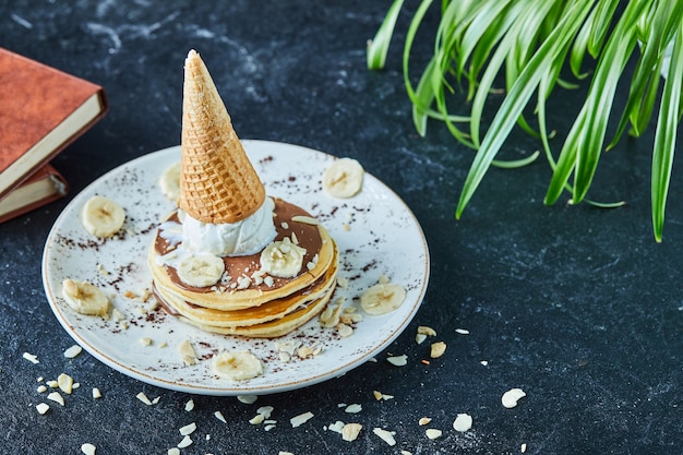 Pancakes with ice-cream cone, bananas, cocoa powder and books in the white plate on the dark surface 