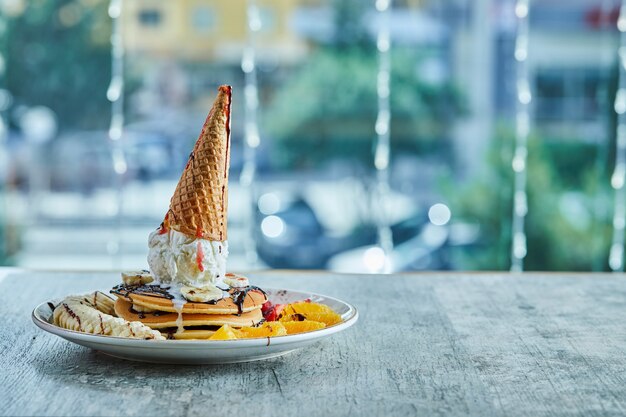 Pancakes with ice-cream cone, tangerine, strawberry, banana and chocolate syrup in the white plate on the marble surface