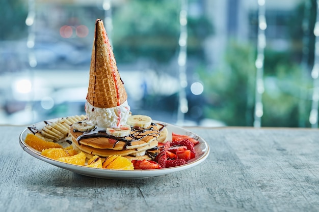 Pancakes with ice-cream cone, tangerine, strawberry, banana and chocolate syrup in the white plate on the marble surface