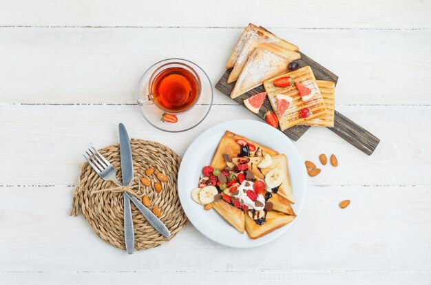 Pancakes on wooden board with tea, almonds, knife, fork, grapes and raspberries