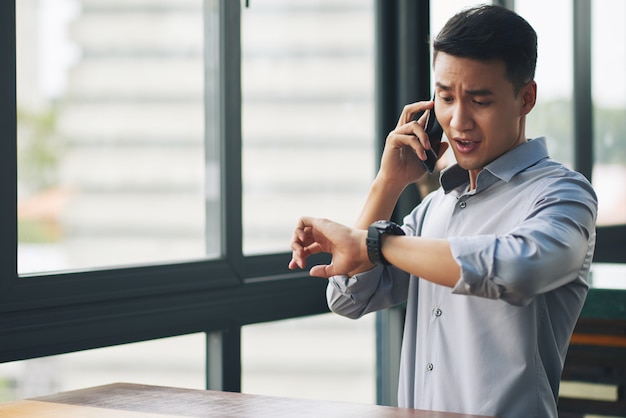 Panicking Asian man talking on mobile phone and looking at wristwatch