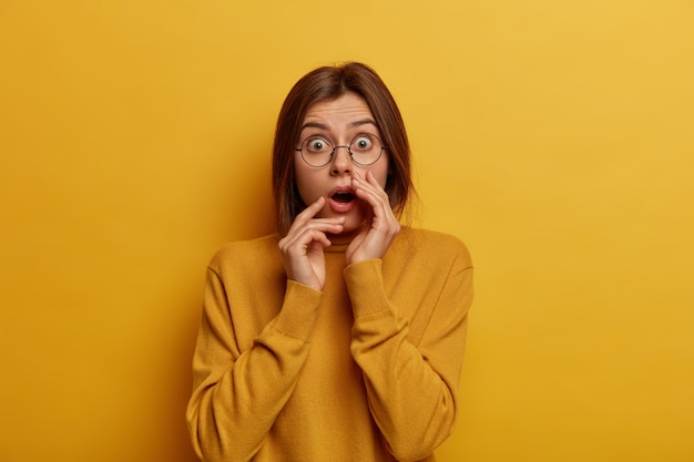 Panicking shocked European woman feels disappointed and impressed, hears something unbelievable, wears round spectacles and casual jumper, isolated over yellow wall, stands speechless.