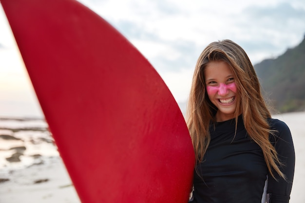 Panoramic view of glad smiling attractive surfer has pink zinc cream applied on face for sun rays protection