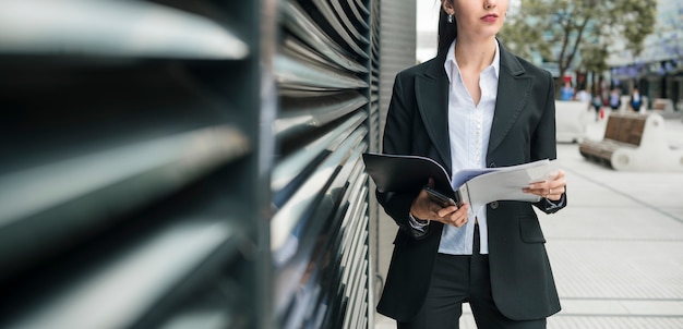 Panoramic view of young businesswoman holding folder and mobile phone in hand standing outside the office