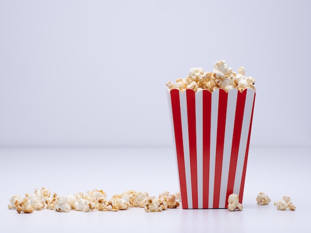 Paper cup of popcorn and some popcorn scattered around on a white surface