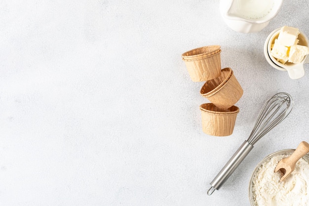 Paper cupcake baking pans on a white concrete background and ingredients Flour butter sugar whisk milk Copy space Top view Baking concept flat lay