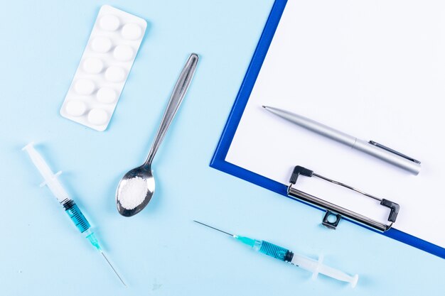 Paper holder and a pen with needles, spoon, pills top view on a light cyan background