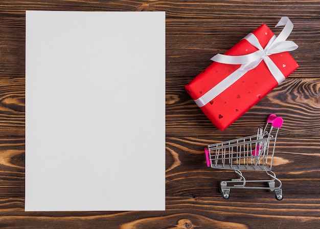 Paper near red present box with white ribbon and shopping trolley