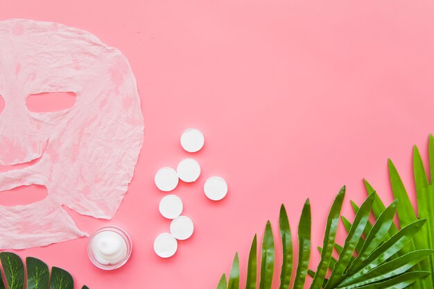 Paper sheet face mask; moisturizer and green leaves on pink background