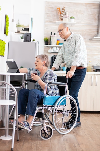 Paralysied senior wife in wheelchair working at tablet computer, showing to husband her project sitting in kitchen. Disabled handicapped old elderly person using modern communication online internet w
