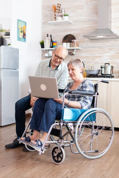 Paralyzed senior woman in wheelchair and husband browsing on internet using laptop in kitchen. Disabled handicapped old elderly person using modern communication techonolgy.