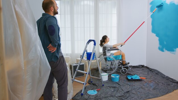 Paralyzed woman painting wall with roller brush sitting on wheelchair. Handicapped, disabled ill and immobilizes woman helping with apartment redecoration and home construction while renovating and im