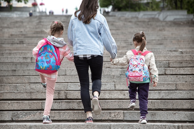 Parent and pupil of primary school go hand in hand. Mom of two girls with a backpack behind the back. Beginning of lessons. First day of fall.