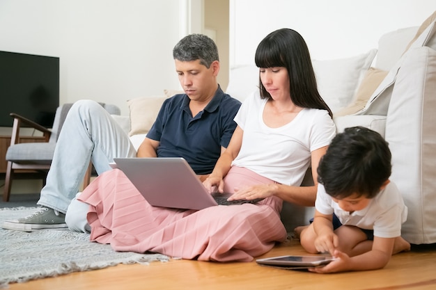 両親のカップルと幼い息子が一緒にアパートの床に座って、タブレットとラップトップのpcを使用しています。