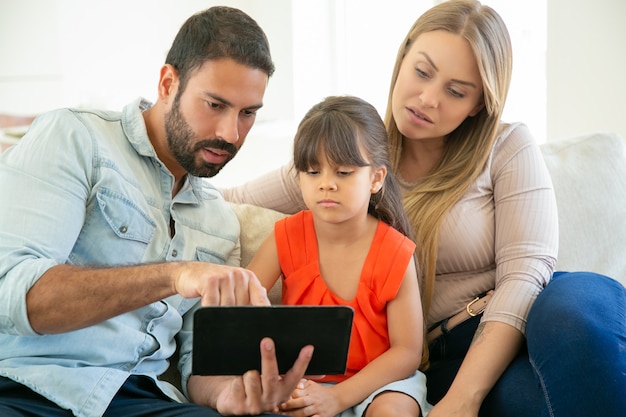 Parents and cute girl sitting on couch, using tablet, watching video together.