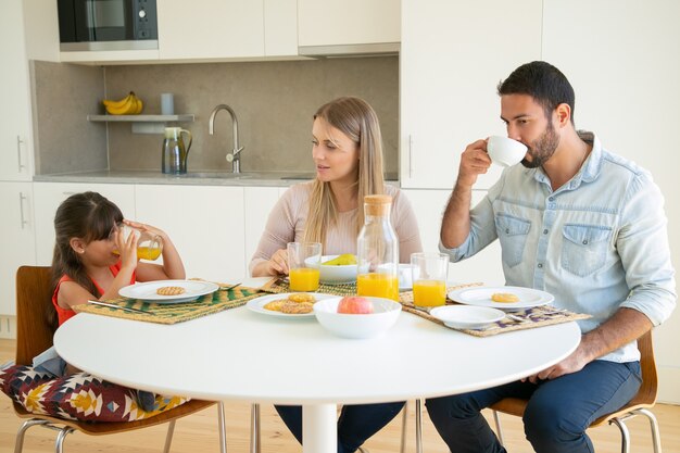 Parents and kid enjoying breakfast together, drinking coffee and orange juice, sitting at dining table with fruits and cookies and talking.