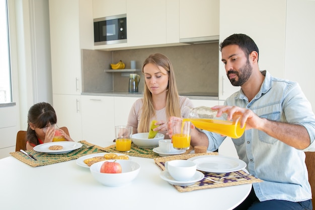 Parents and kid sitting at dining table with dish, fruit and cookies, pouring and drinking fresh orange juice.