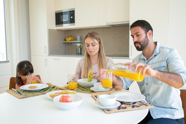 Parents and kid sitting at dining table with dish, fruit and cookies, pouring and drinking fresh orange juice.