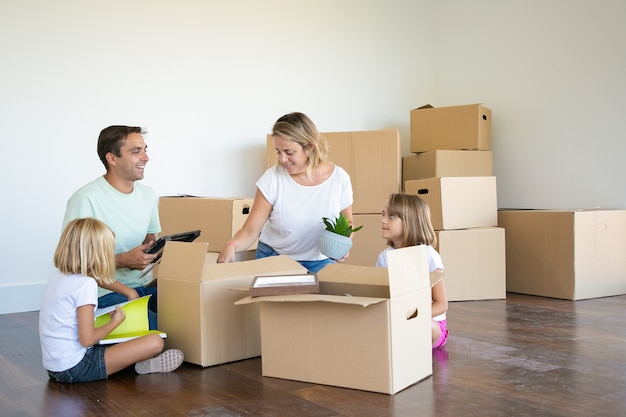 Parents and kids unpacking things in new apartment, sitting on floor and taking objects from box