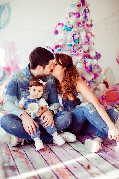 Родители целуются с маленькой дочерью перед яркой рождественской елкой