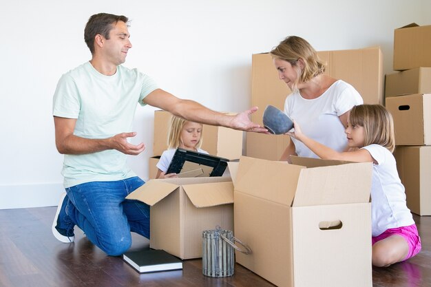 Parents and little daughters unpacking things in new apartment, sitting on floor and taking objects from open boxes