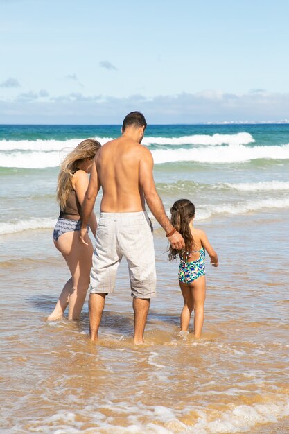 Parents and little girl in swimsuits walking ankle deep in ocean waves