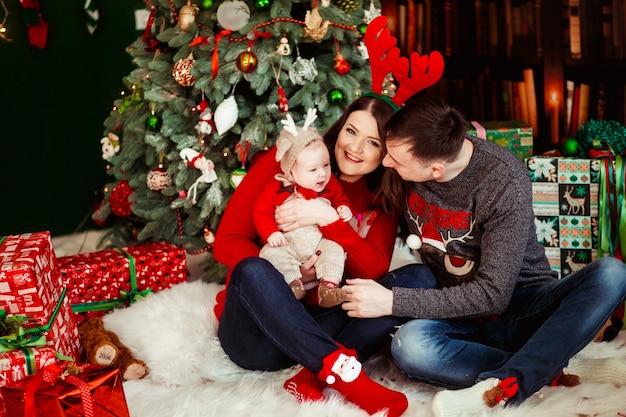 Parents play with little daughter in deer hat holding her on their arms and sitting before a Christmas tree