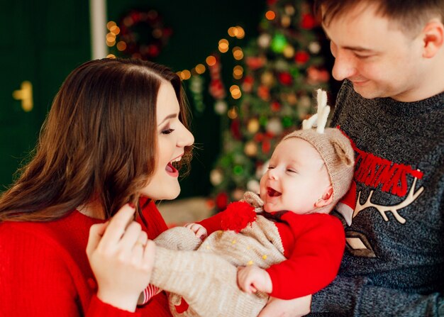 부모는 사슴 모자에 작은 딸과 함께 노는 그녀의 팔에 그녀를 잡고 크리스마스 트리 앞에 서