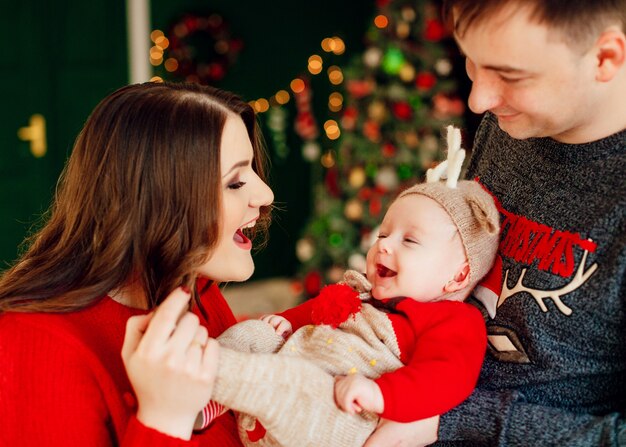 Parents play with little daughter in deer hat holding her on their arms and standing before a Christmas tree