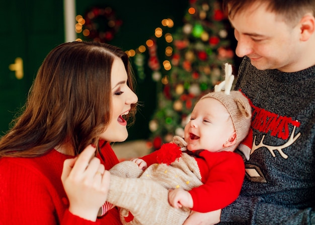 親は彼女を腕につけてクリスマスツリーの前に立っている鹿の帽子で小さな娘と遊びます