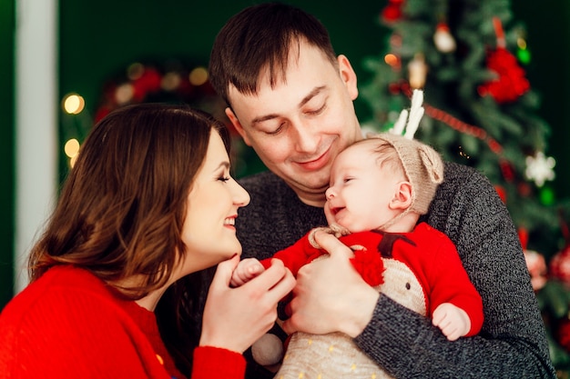 Parents play with little daughter in deer hat holding her on their arms and standing before a Christmas tree