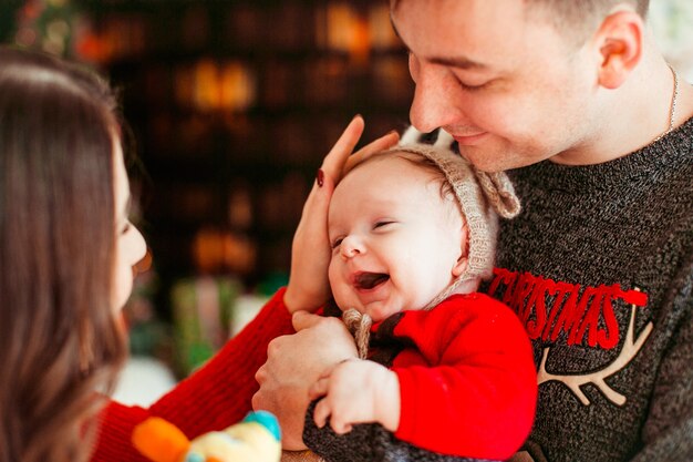 Родители играют с маленькой дочкой в ​​шляпе оленя, держа ее на руках и стоят перед рождественской елкой