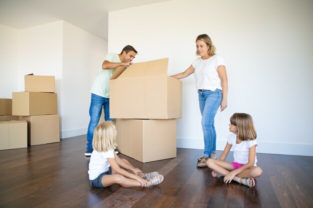 Родители и две дочери открывают коробки и распаковывают вещи в своей новой пустой квартире.