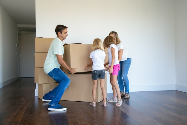 Parents and two girls carrying boxes and making stack carefully in their new empty flat