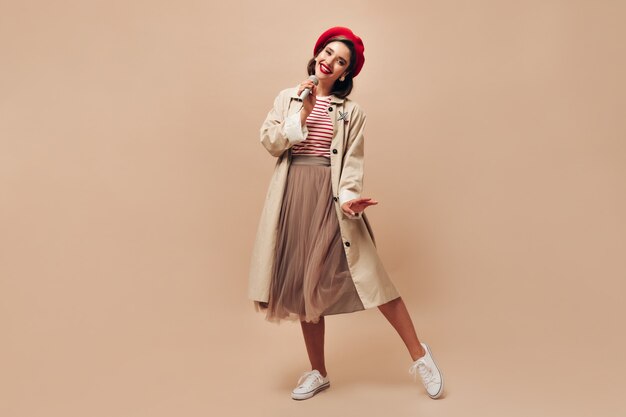 Parisian lady in beret and trench sings in microphone. Stylish brunette in long skirt and light autumn coat posing on camera.