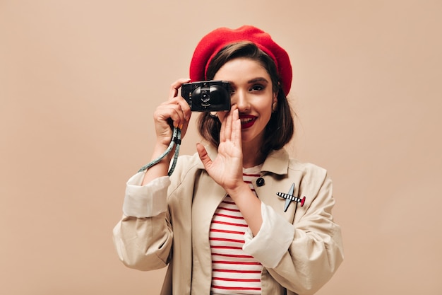 Parisian woman in beret takes photos on beige background. Cute young girl with dark hair and bright lips in autumn coat holds black camera.