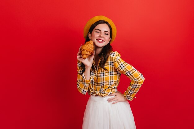 Parisian woman in stylish yellow shirt holding appetizing croissant. Lady in beret with smile on red wall.