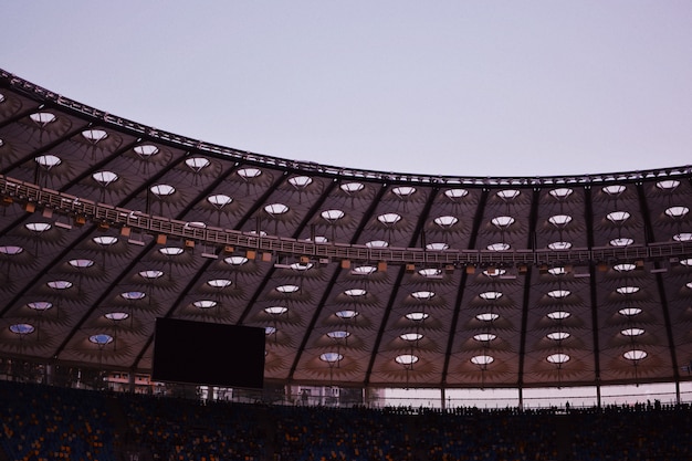 Partial shot of a stadium displaying the roof, a large monitor top seating rows and chairs